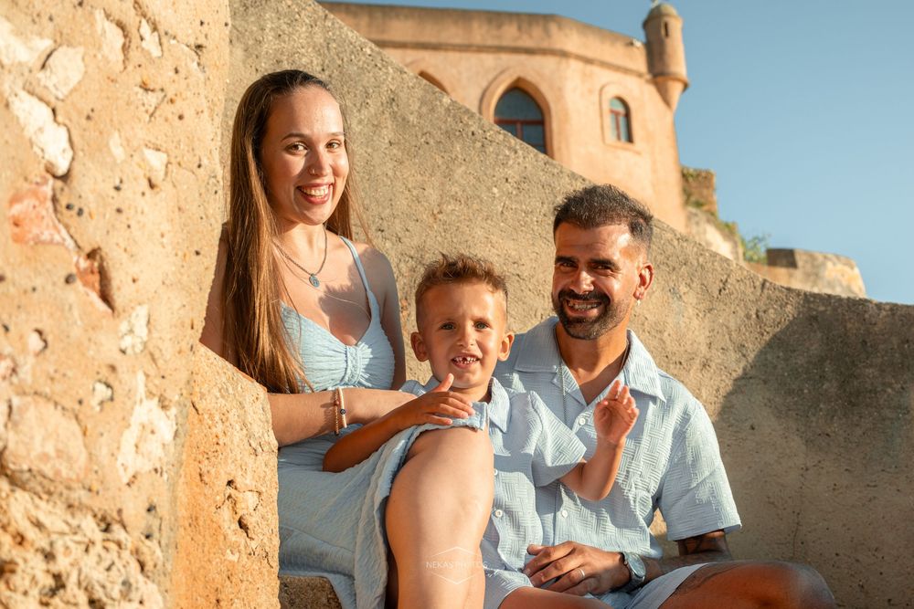 Happy family of three posing near ancient stone walls in warm golden sunlight at a historic Mediterranean landmark.