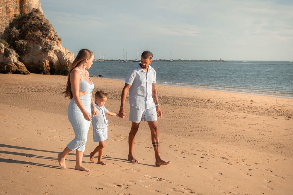 Family of three walking hand-in-hand on a sunny sandy beach with rocky cliffs and calm blue ocean.
