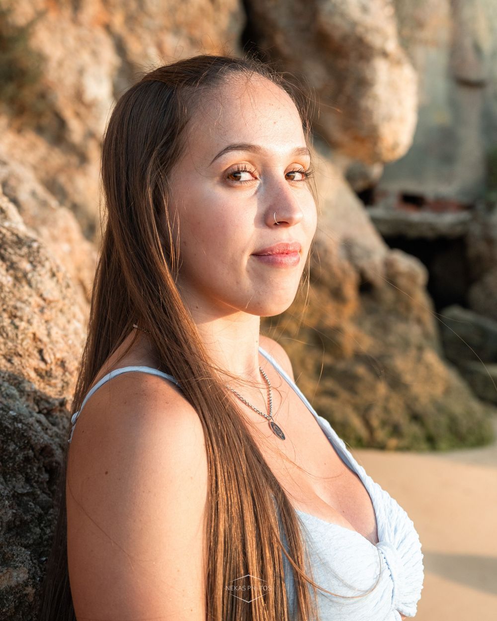 Young woman with long brown hair poses near rocky beach cliffs in golden hour sunlight wearing a white top.