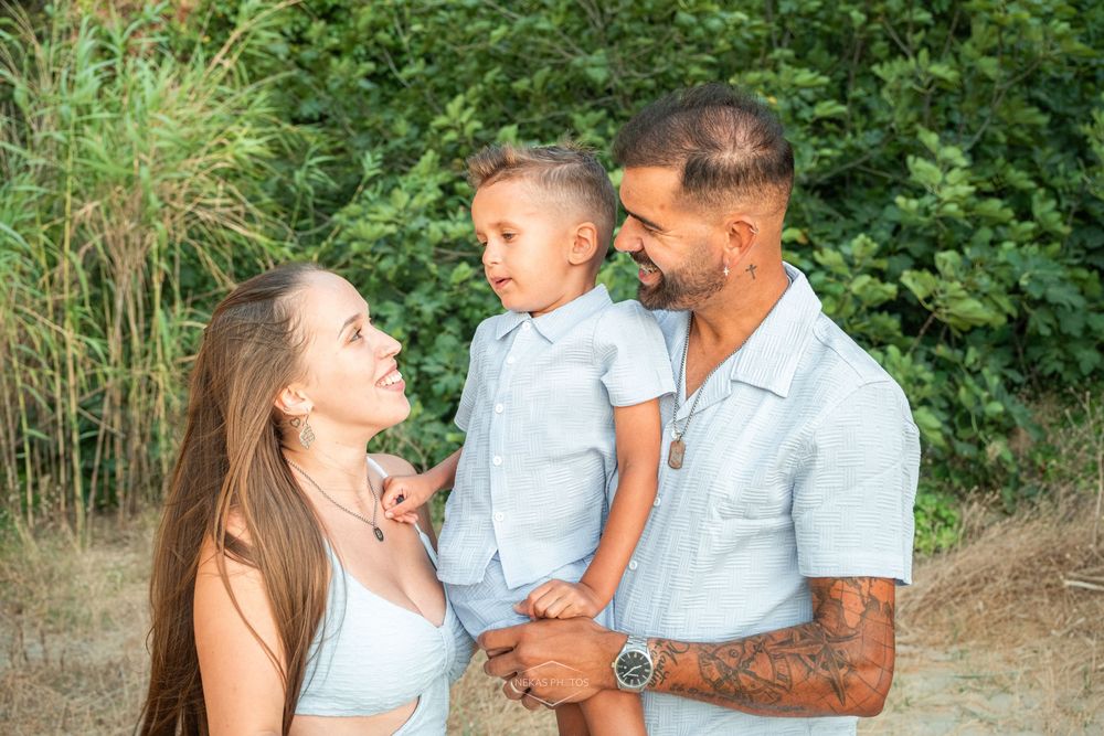 A young family with a tattooed father holding a toddler boy, smiling with mother outdoors near green trees.