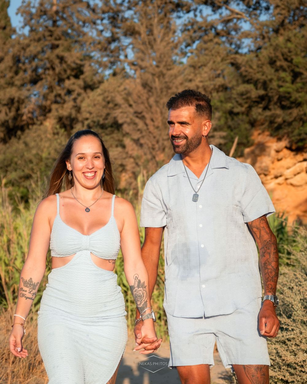 A smiling couple holding hands and walking on a sunny path, both wearing matching light blue outfits with visible tattoos.
