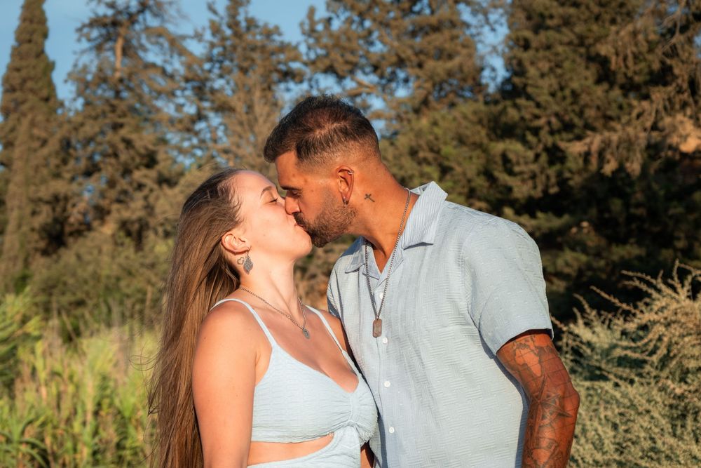 A couple shares a romantic kiss outdoors surrounded by lush green trees in warm golden sunlight.