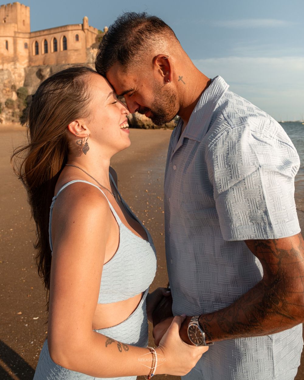A couple shares a romantic moment on a beach at golden hour with a historic building in the background.