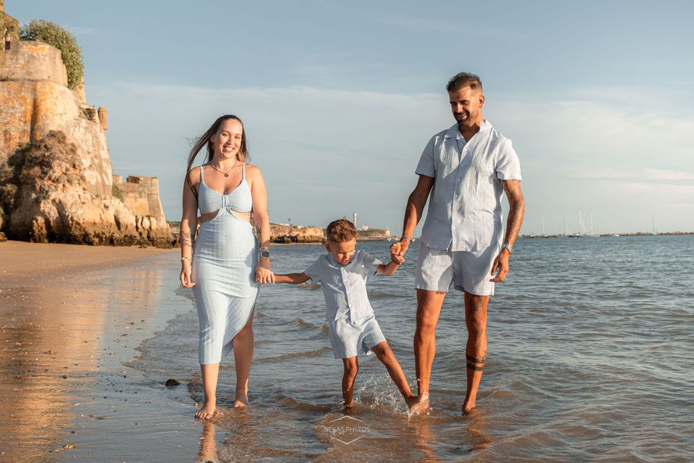 Pregnant woman and man hold child's hands walking on sunny beach in white outfits near rocky cliffs.