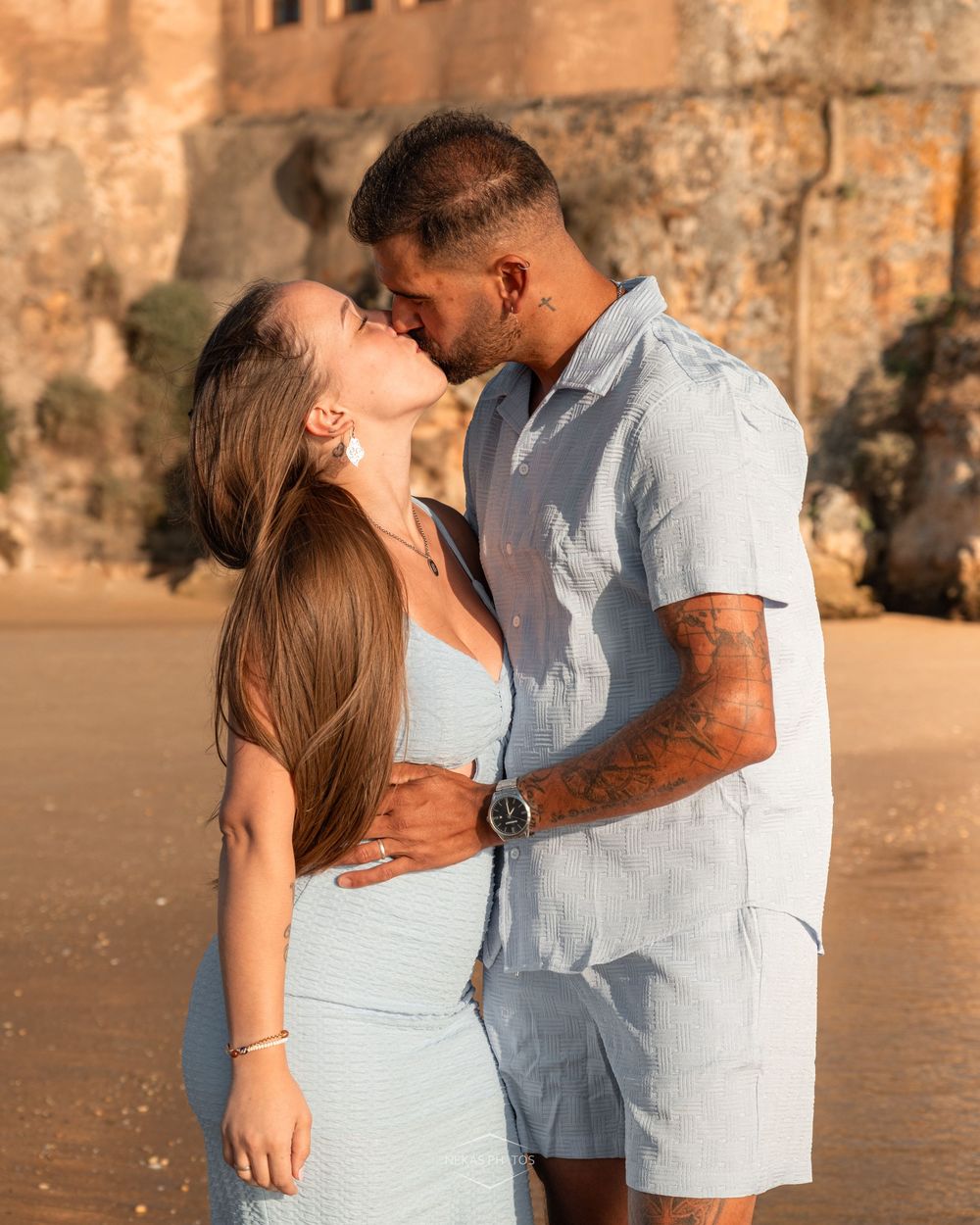 Couple sharing a romantic kiss on a beach at golden hour, wearing matching light blue outfits near ancient stone ruins.