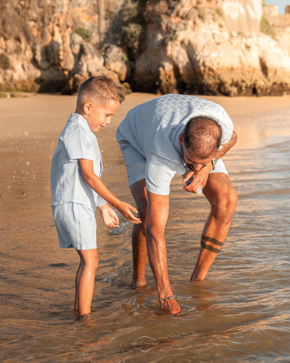 A man and young boy in matching light blue outfits explore the shallow ocean waves together on a sandy beach.