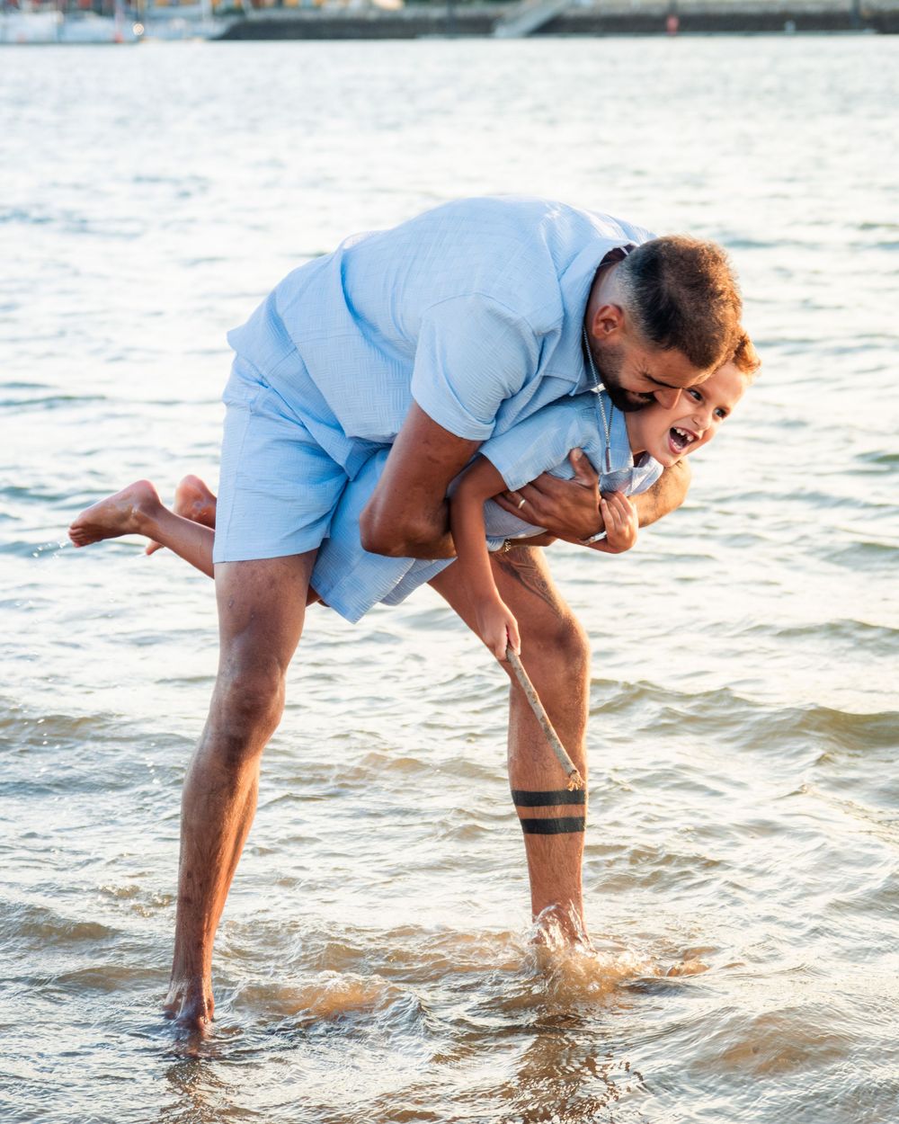 A man playfully carries a laughing child over his shoulder while standing in shallow water at the beach.