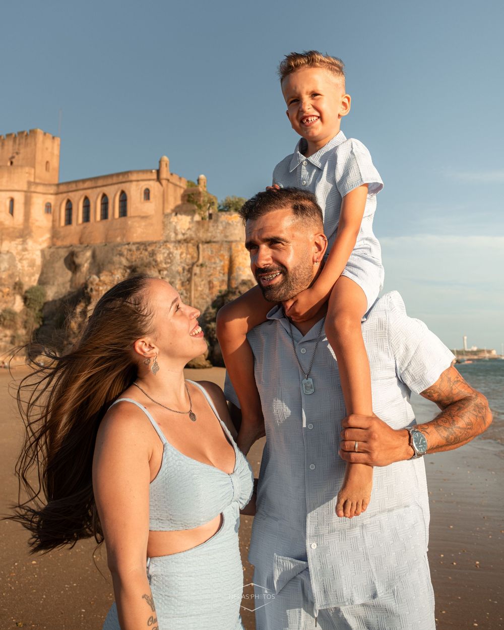Happy family of three on a sunny beach, parents smiling at each other with young boy sitting on dad's shoulders near ancient ruins.