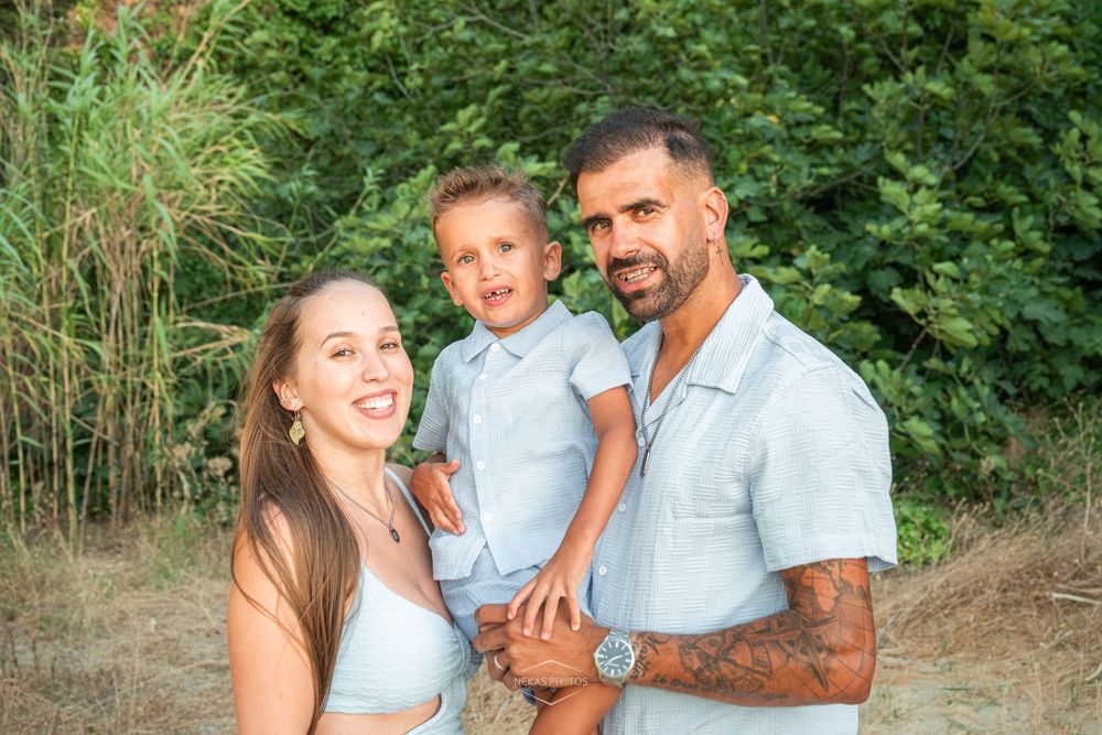 A smiling family of three poses outdoors, with a tattooed father holding a young boy beside a happy mother near green trees.