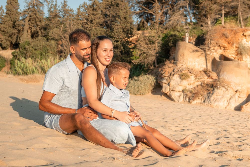 Family of three sitting on sandy beach at golden hour, with rocky cliffs and trees in background.