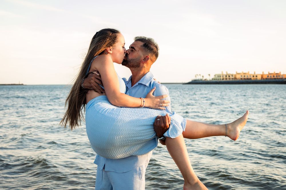 Romantic couple kissing by the sea, man lifting woman in his arms at sunset near a waterfront.
