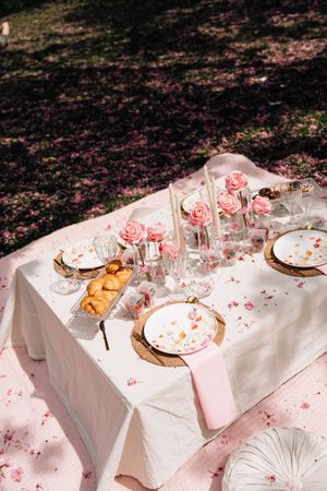 Elegant outdoor picnic table with pink roses, gold chargers, and croissants on a white linen under cherry blossom trees.