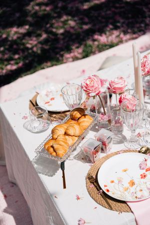 Elegant outdoor brunch table with croissants, floral plates, pink roses, crystal glasses, and candles on a white tablecloth.