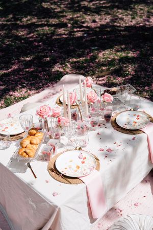Elegant outdoor garden table setting with pink roses, white candles, floral plates, and croissants on a white linen tablecloth.