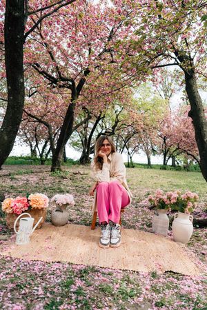 Woman in pink pants sits on chair under blooming cherry blossom trees surrounded by floral arrangements on a petal-covered rug.
