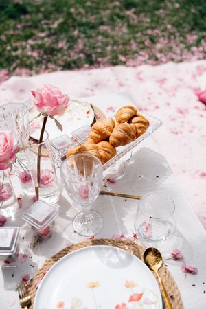 Elegant outdoor picnic setup with croissants, crystal glasses, pink roses, and gold cutlery on a white cloth with scattered petals.