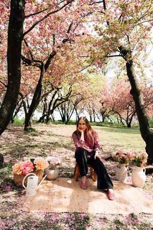 A young woman sits among pink cherry blossom trees surrounded by flower arrangements in a serene spring garden setting.