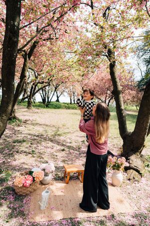 A mother lifts her child under blooming pink cherry blossom trees on a picnic blanket with flowers.