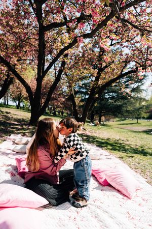 Mother and child sharing a kiss on a picnic blanket under blooming pink cherry blossom trees in a park.
