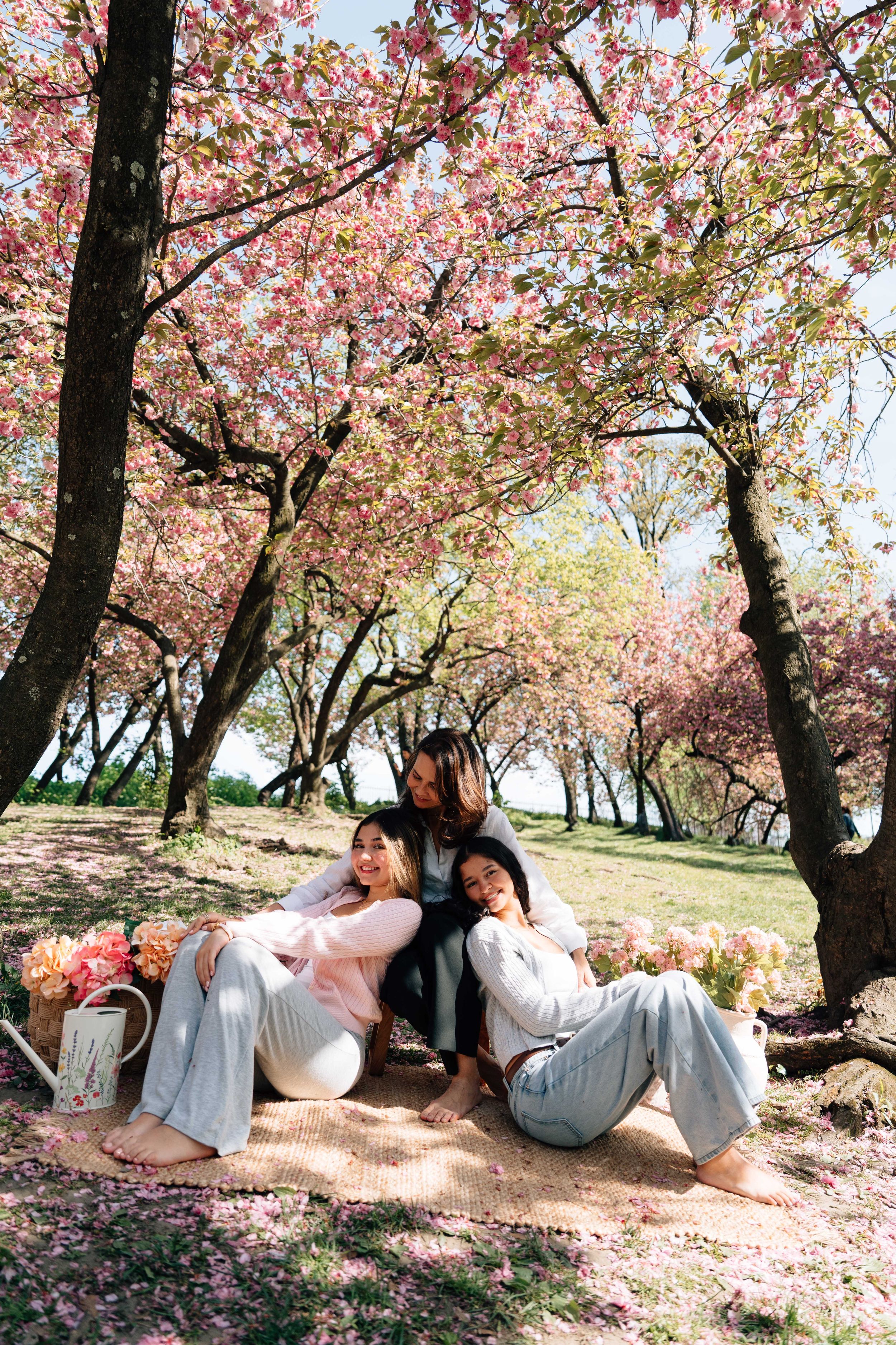 Three women relaxing on a blanket under blooming pink cherry blossom trees in a sunny park setting.