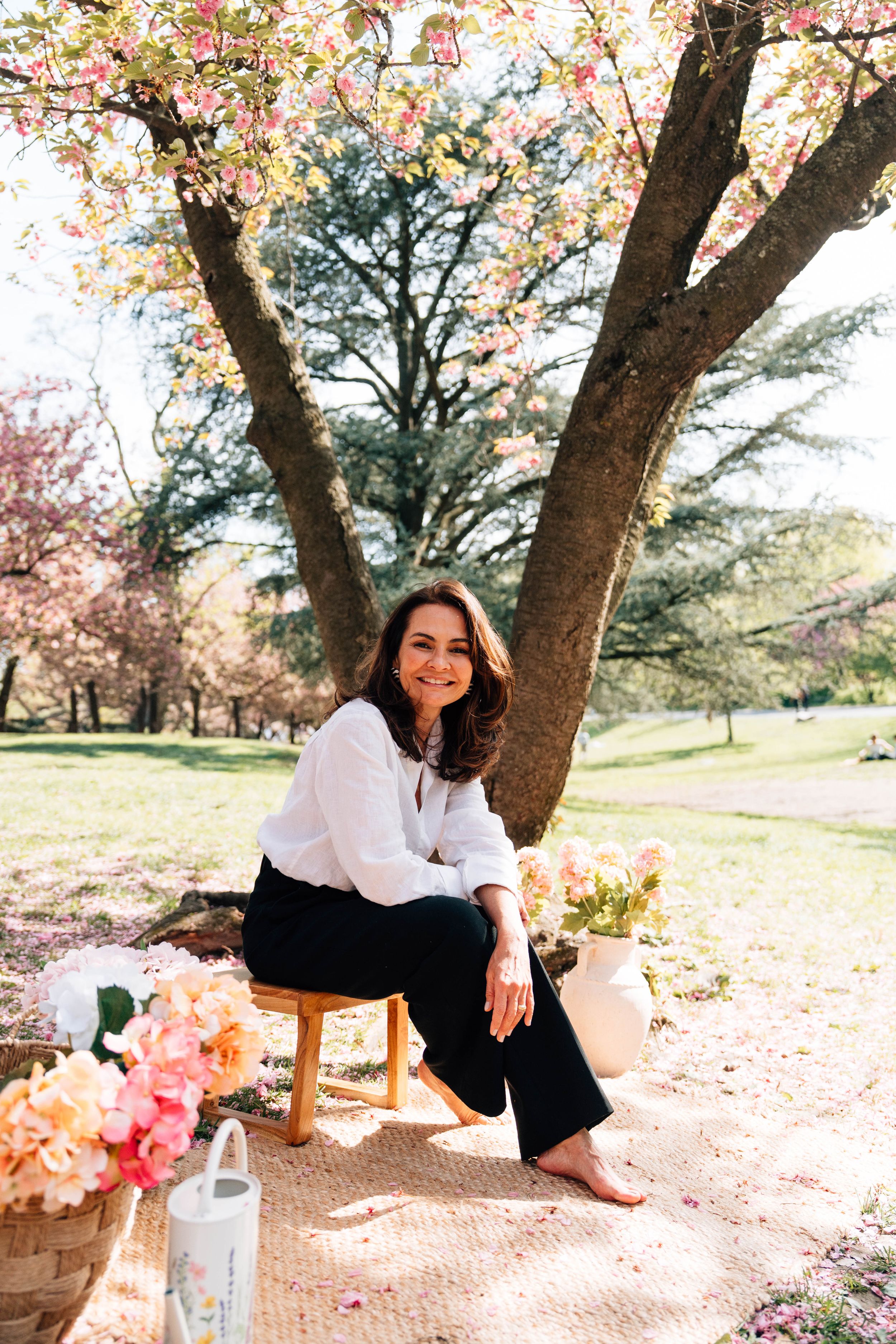 Woman sitting on stool under blooming cherry blossom tree in spring park with pink flowers and vase nearby.