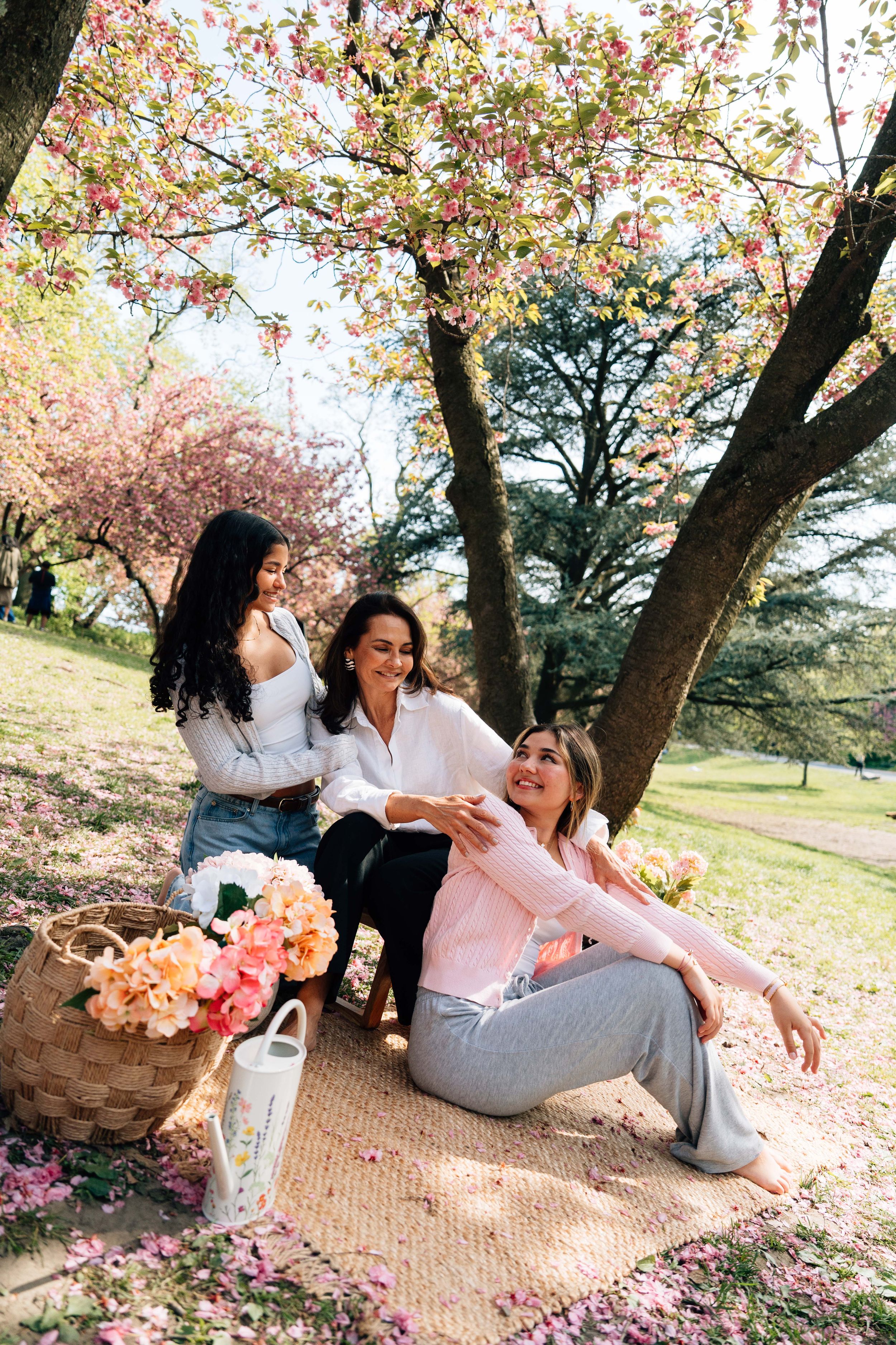 Three women enjoying a spring picnic under blooming cherry blossom trees in a park with flowers.