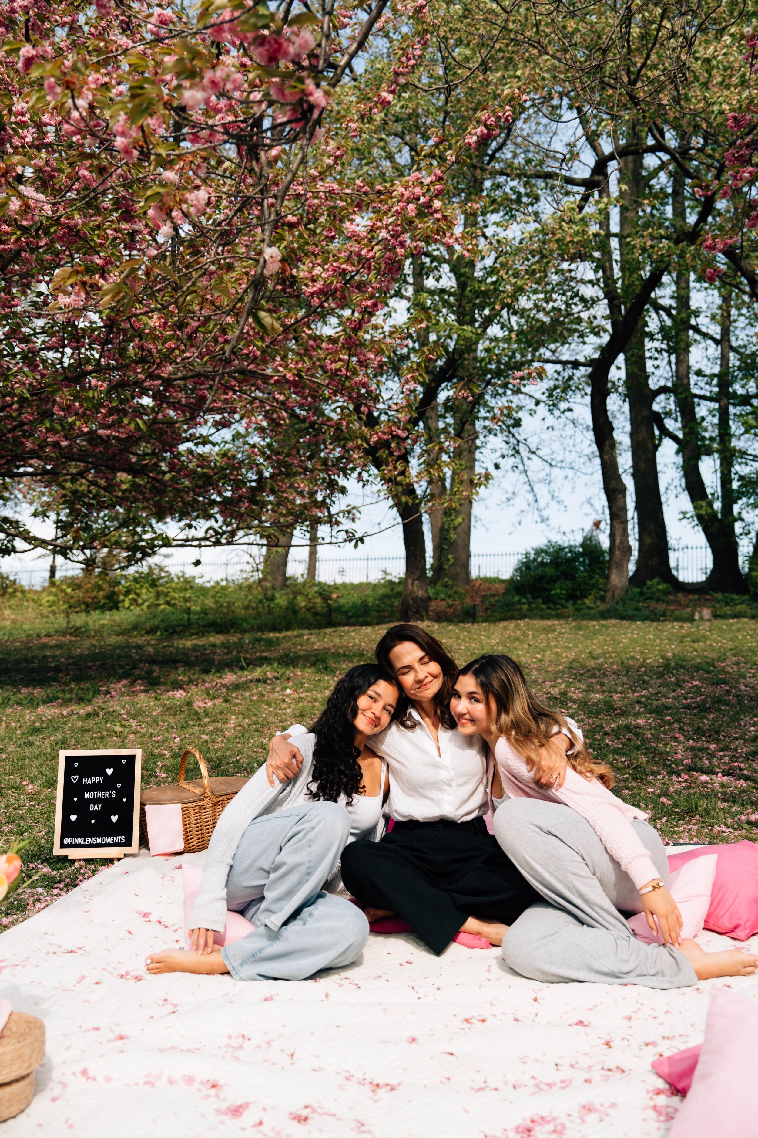 Three women sitting together on a blanket under blooming cherry blossom trees during a spring picnic.