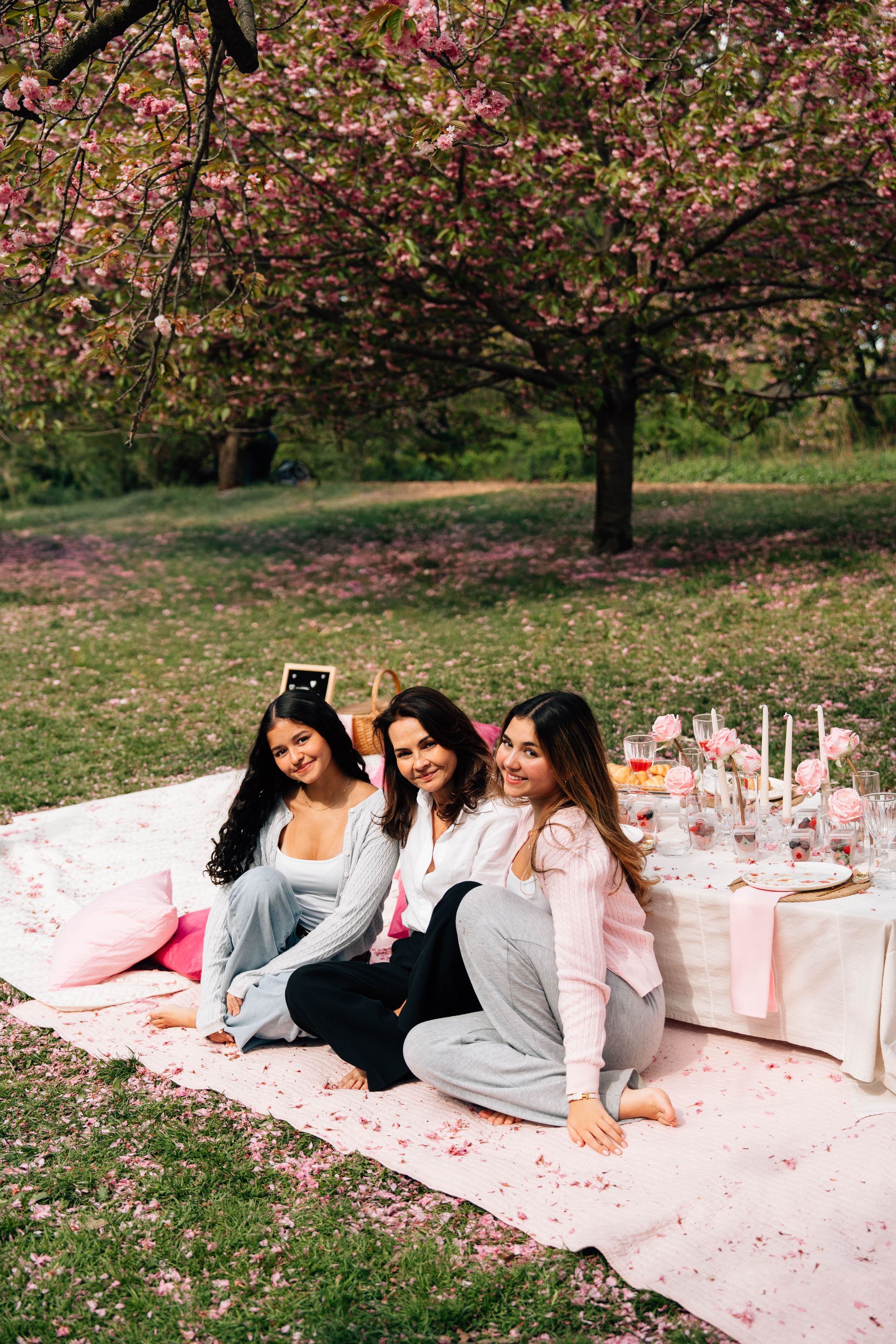Three women sitting on a blanket taking a selfie under blooming pink cherry blossom trees at a spring picnic.