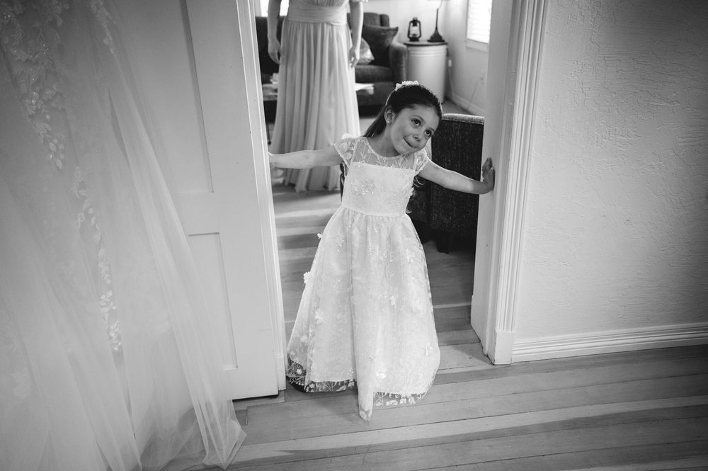 Black and white photo of a young flower girl in a lace dress smiling in a doorway at a wedding.