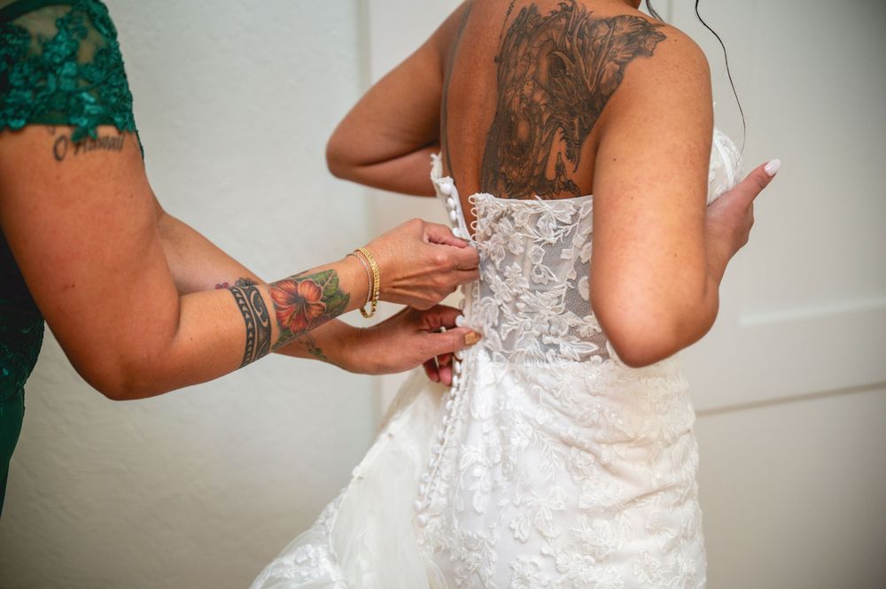 Tattooed bridesmaid lacing up a white lace wedding dress on a bride from behind.