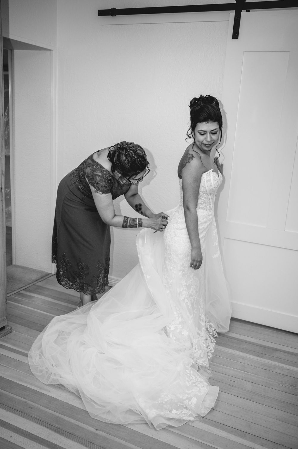 Black and white photo of a bride in a lace gown being helped by a woman adjusting her dress before the wedding ceremony.