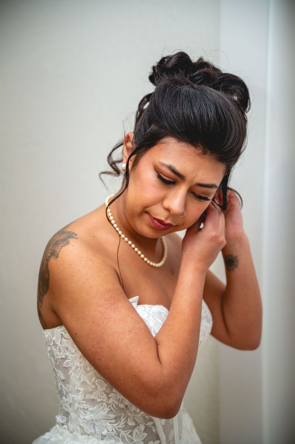Bride in strapless lace gown adjusting her earring, wearing pearl necklace with elegant updo hairstyle.