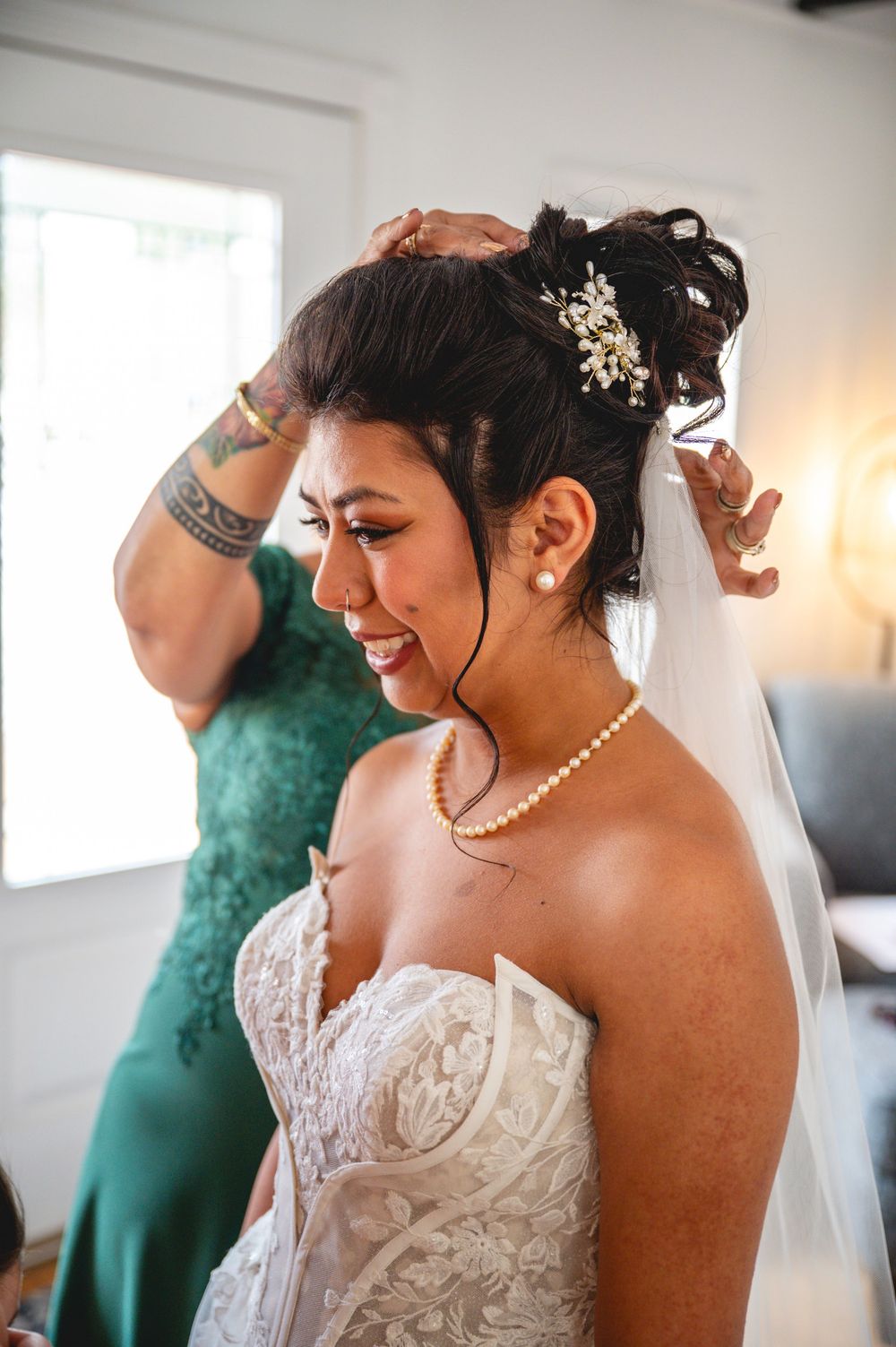 Bride in strapless lace gown with pearl necklace and veil gets hair styled before her wedding ceremony.