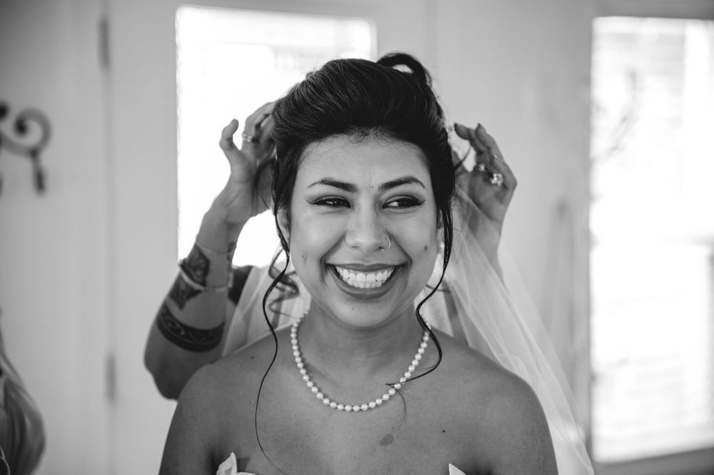 Smiling bride in black and white photo wearing pearl necklace and veil being adjusted on wedding day.