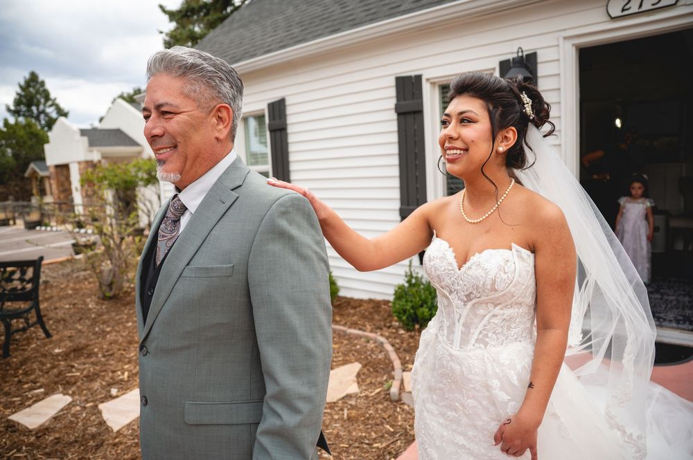 Bride in strapless lace gown smiles at her father in gray suit outside a white chapel on wedding day.