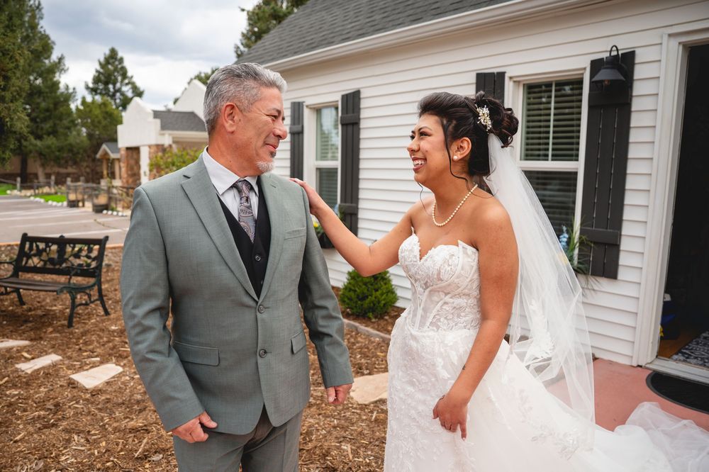 Bride in white gown and groom in gray suit share a joyful first look moment outside a white building.