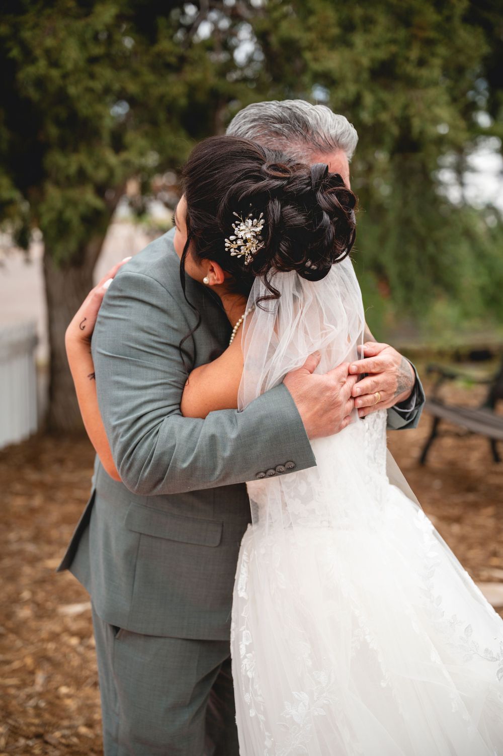 A bride and older man share an emotional hug outdoors, her lace gown and jeweled updo visible in a tender wedding moment.
