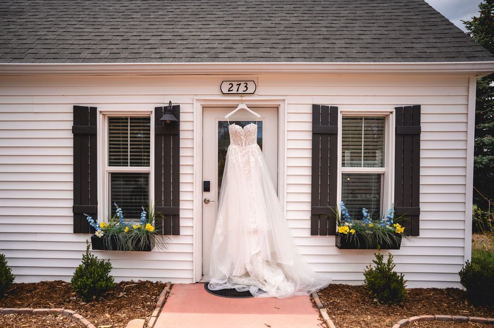 A white ball gown wedding dress hanging on the front door of a charming white cottage with black shutters.