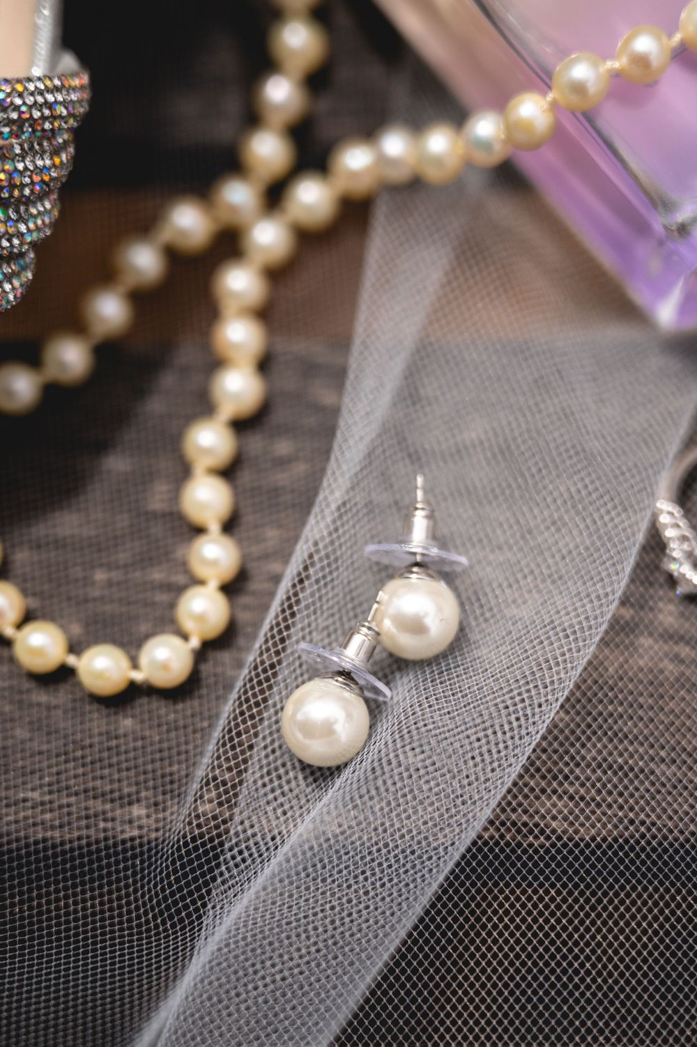 Pearl earrings and necklace displayed on a wedding veil with a purple perfume bottle and rhinestone accessories.