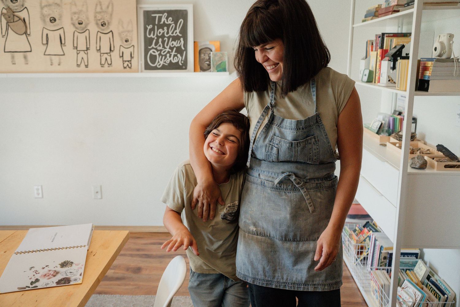 A woman in a denim apron smiles with a young girl leaning on her in a bright, art-filled studio space.