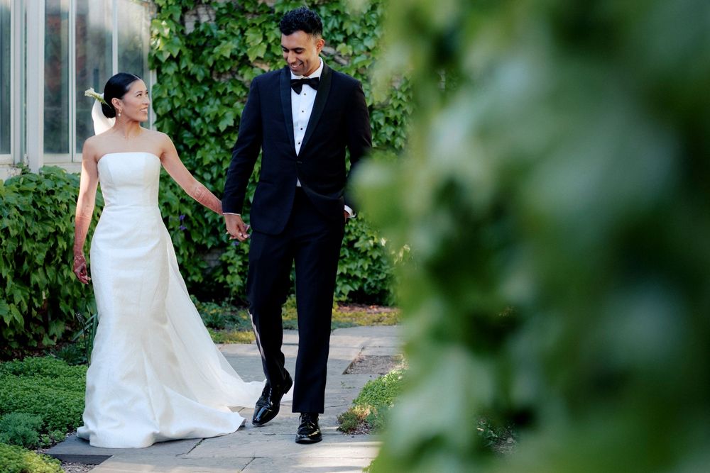 Filipino bride and Indian groom walk hand in hand through a lush garden pathway, bride in strapless white gown, groom in black tuxedo.