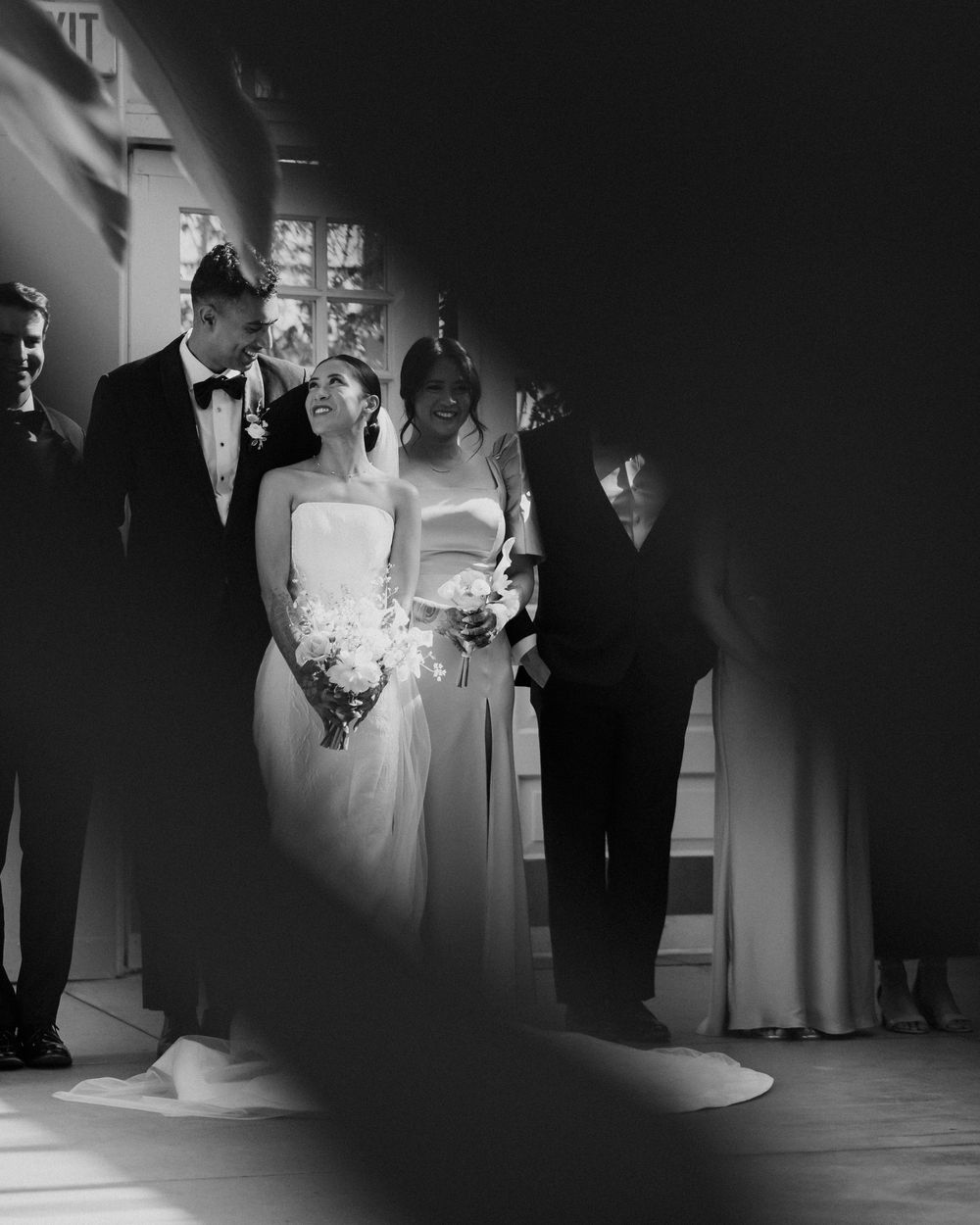 Artistic film wedding photo of wedding party smiling, bride in elegant gown holding bouquet, shot through foliage.