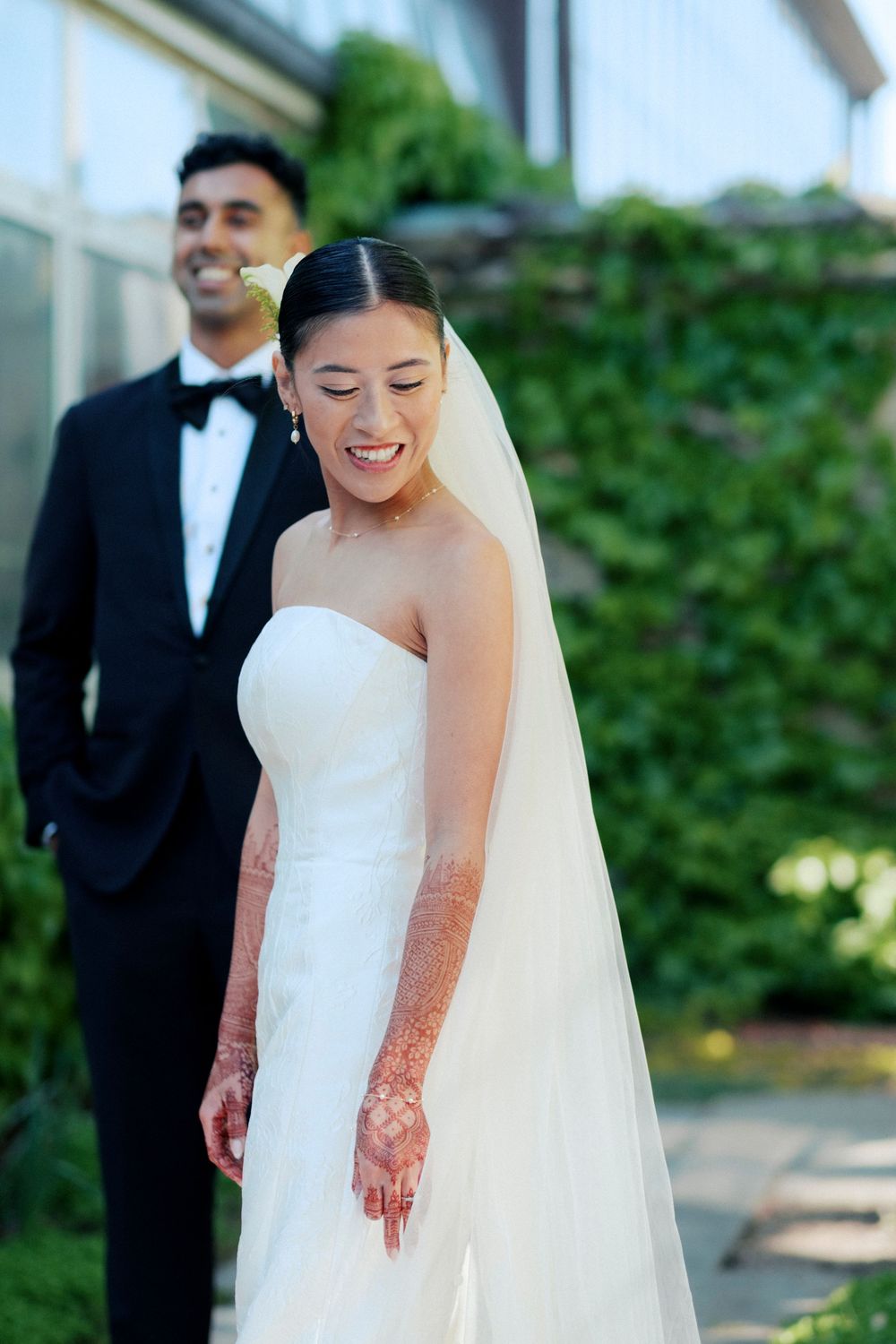 Smiling Asian bride in white strapless gown with henna and veil, groom in black tuxedo behind her outside Garfield Conservatory.
