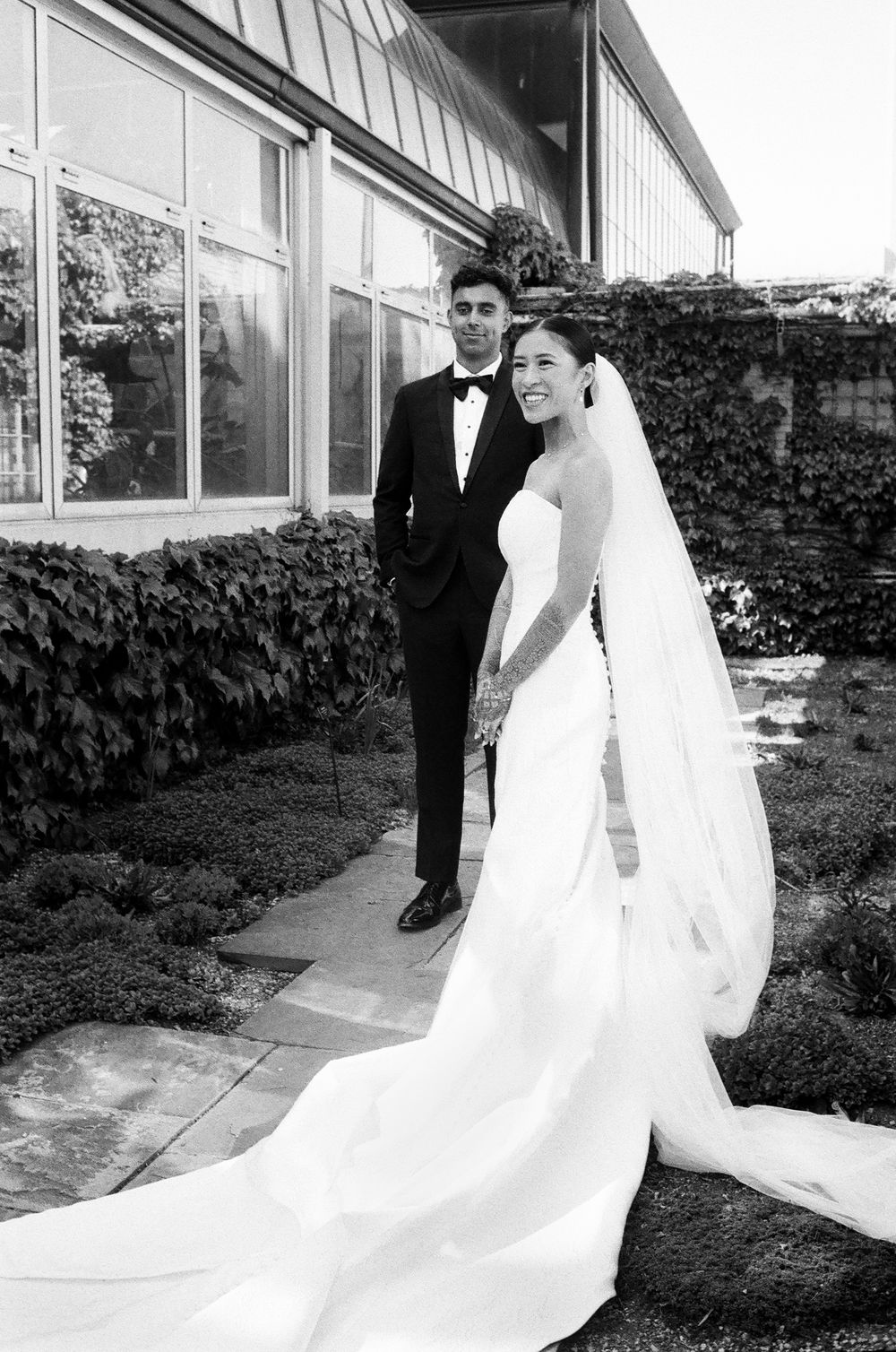 Black and white film wedding photo of bride in strapless gown with veil and groom in tuxedo posing outside elegant Midwest venue.