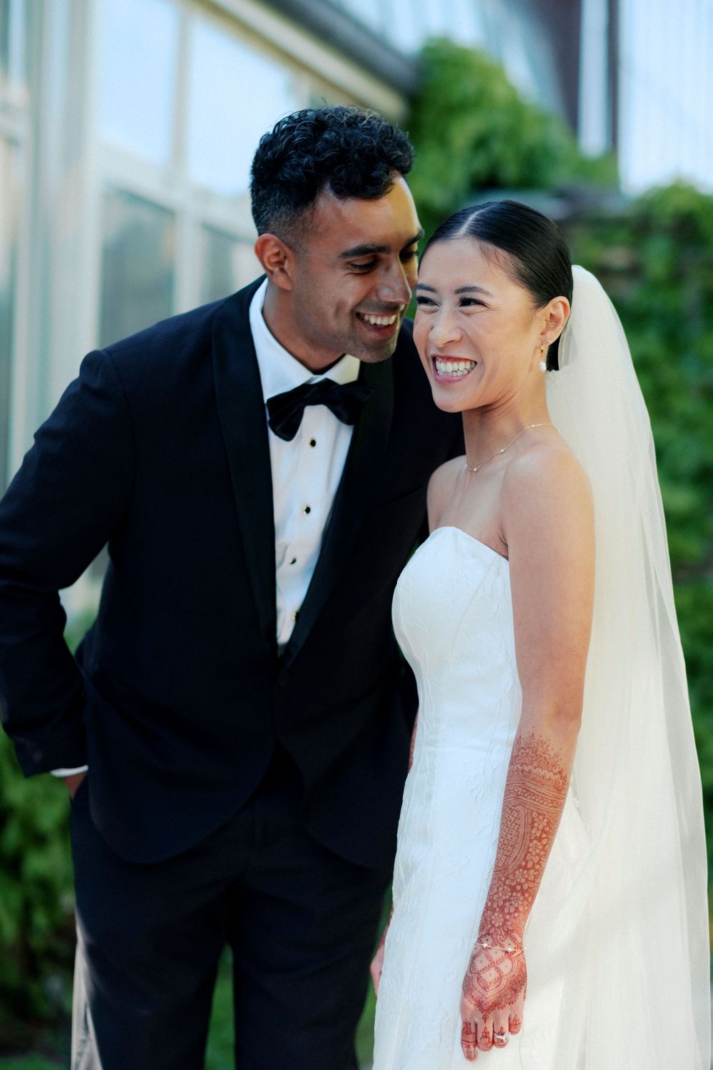 A joyful interracial couple in wedding attire, groom in tuxedo and bride in white gown with veil and henna, smiling outdoors.