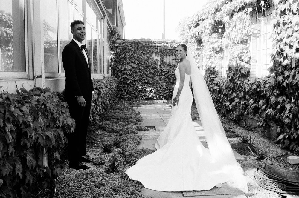 Candid wedding photo on film of bride in elegant gown with veil and groom in tuxedo standing in ivy-covered garden during their first look.