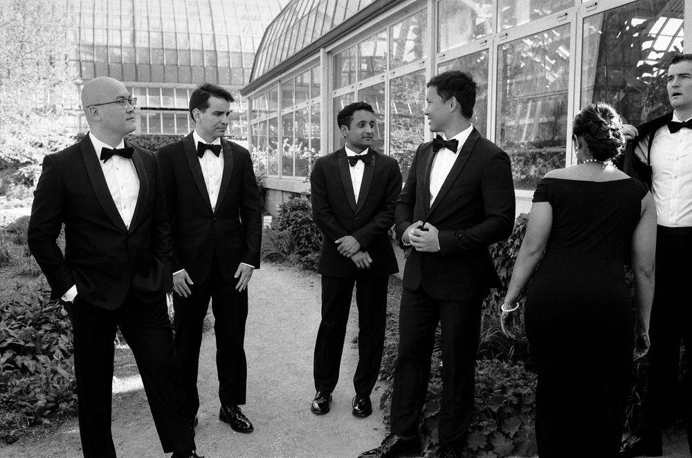 Black and white photo of groomsmen in tuxedos conversing outdoors near a greenhouse at a wedding.