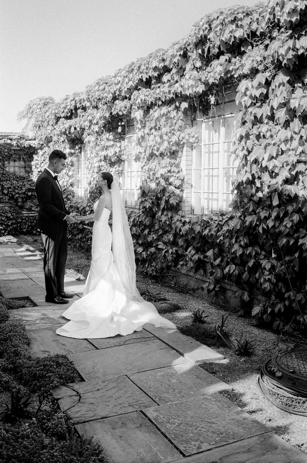 Black and white photo on 35mm film of a bride and groom standing on a stone path beside an ivy-covered building on their wedding day.