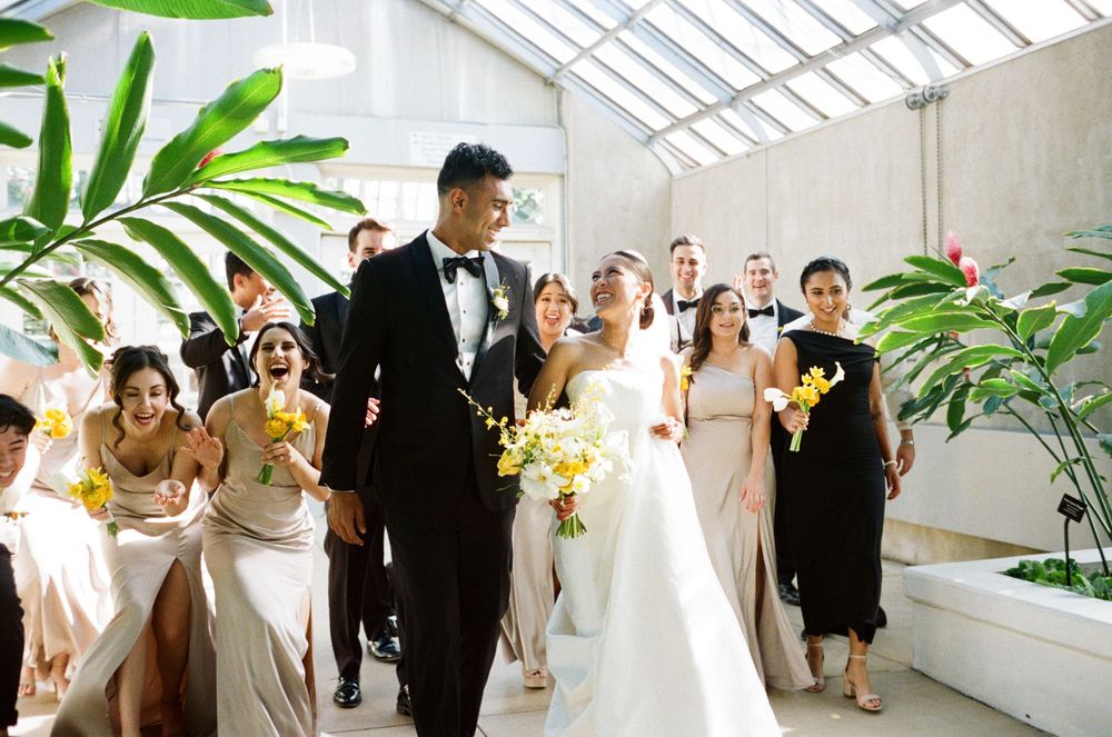 Bride and groom walk joyfully through greenhouse venue aisle surrounded by bridesmaids in champagne gowns holding yellow bouquets.