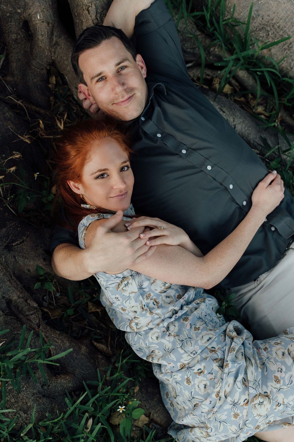 A couple lies together on grass beneath a tree, the woman in a floral dress resting on the man in a dark shirt.